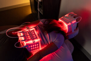 Client's arm and hand wrapped in glowing red LED light therapy pads in a dim treatment room